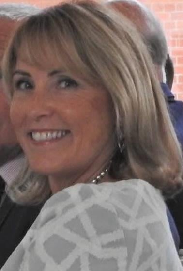 Woman with shoulder-length blonde hair and bangs smiling at camera, wearing a patterned white top and earrings