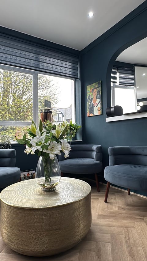 Modern living room with dark teal walls, black armchairs, round gold coffee table with white flowers, and large windows overlooking trees
