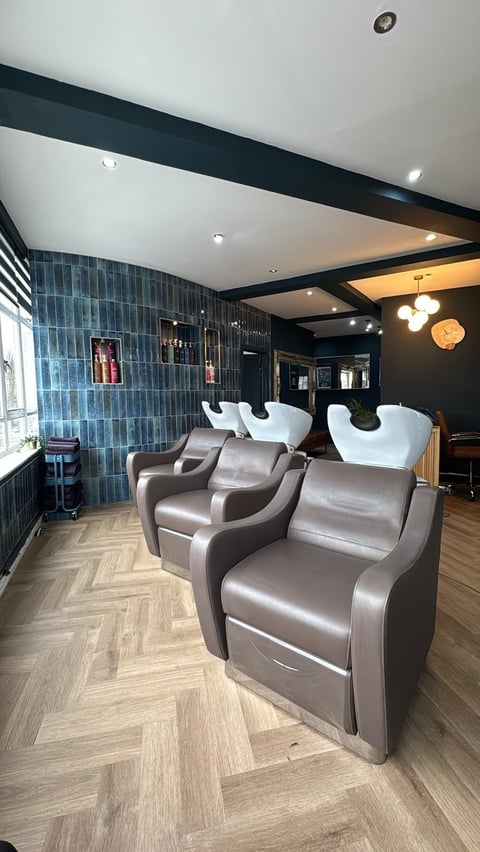 Modern salon interior with gray leather shampoo chairs, blue tiled wall, white ceiling, and herringbone wood flooring