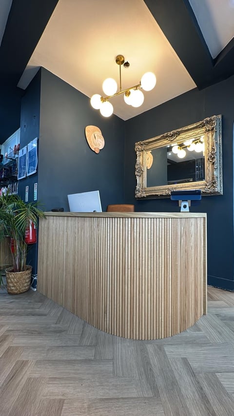 Modern reception desk with vertical wood slats, dark teal walls, gold-framed mirror, and brass globe chandelier