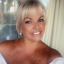 Smiling woman with short blonde hair and bangs, wearing a white top, photographed indoors near a window