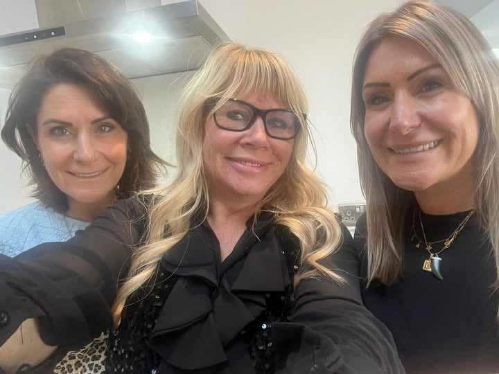 Three smiling women posing together for a selfie indoors, wearing casual black clothing