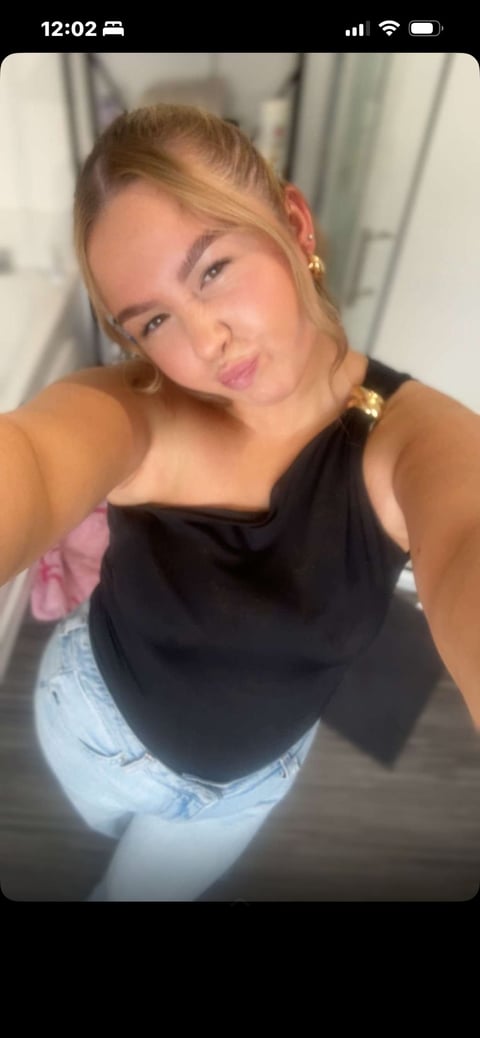 Woman taking a selfie indoors wearing a black sleeveless top and light blue jeans, with gold earrings, smiling at camera